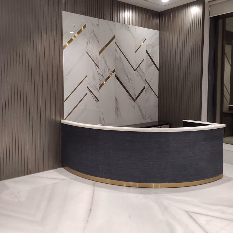 Curved corporate reception desk in dark textured cladding with a white stone counter top and a brass base line, standing in front of a white marble wall inlaid with brass chevron lines.