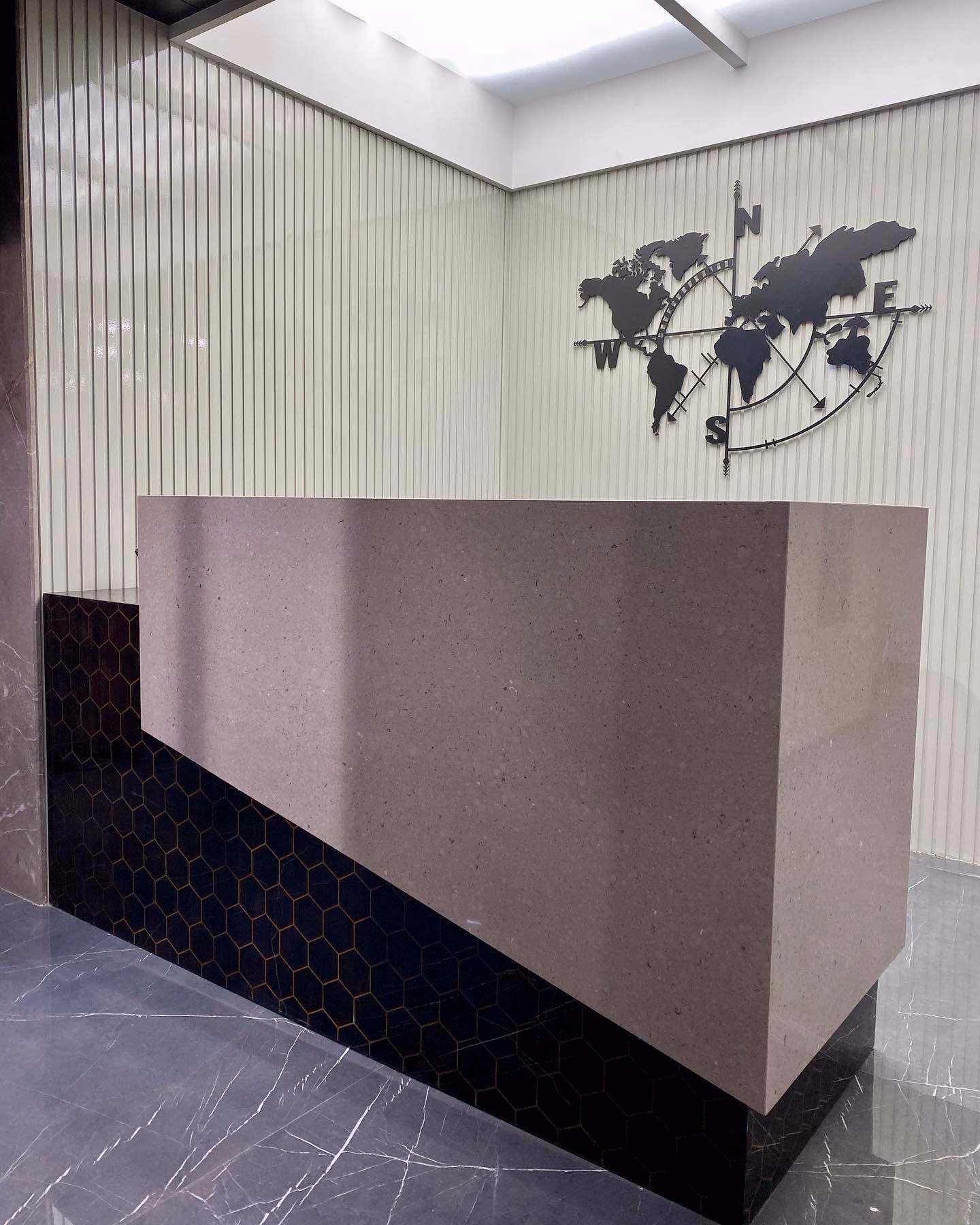 Rectangular reception desk built as a solid stone block with mitred corners, sitting on a recessed black hexagon-tile plinth, against a fluted cream wall with a black world-map compass sculpture.