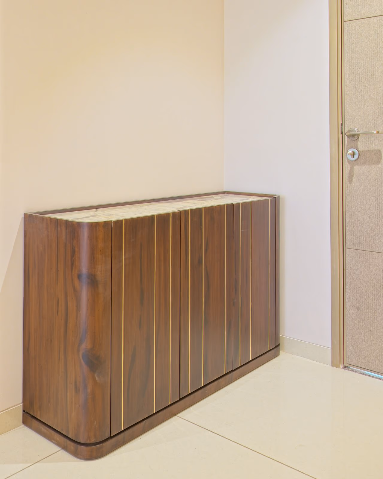 Tall walnut shoe cabinet with vertical brass inlay stripes and a white marble top beside an entry door.