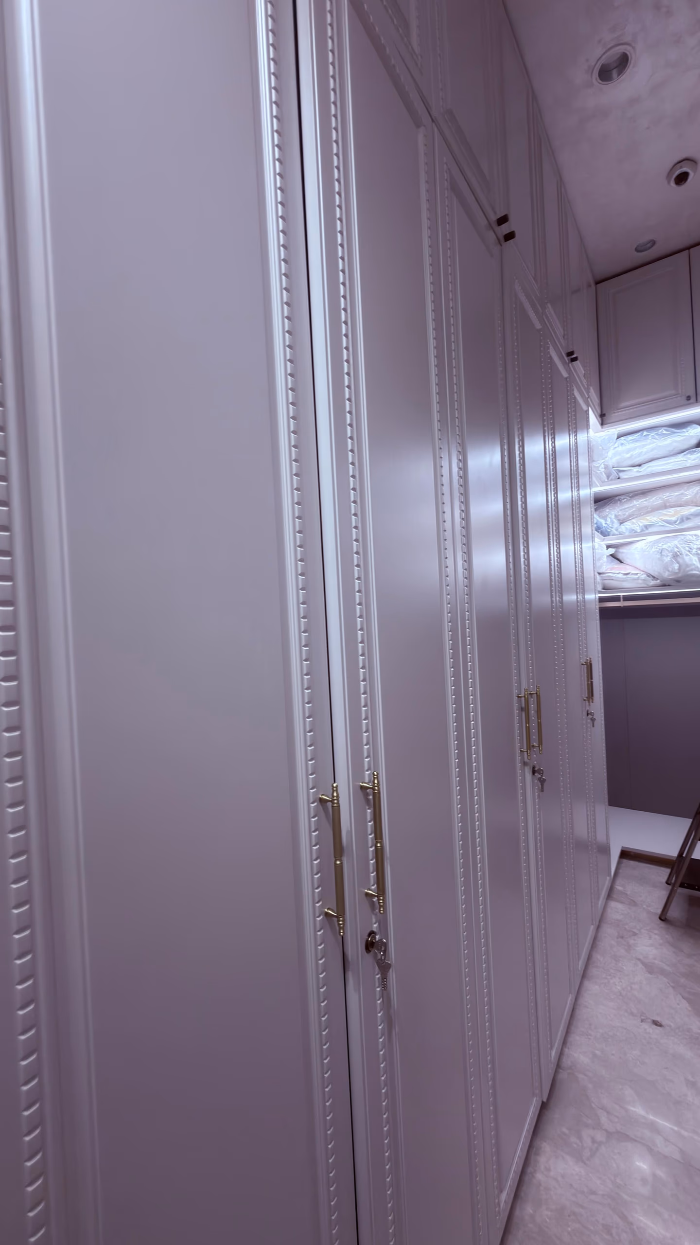 Run of tall soft-lilac lacquer wardrobes reaching the ceiling, edged with beaded moulding trim and slender brass bar handles, lining a marble-floored dressing room.