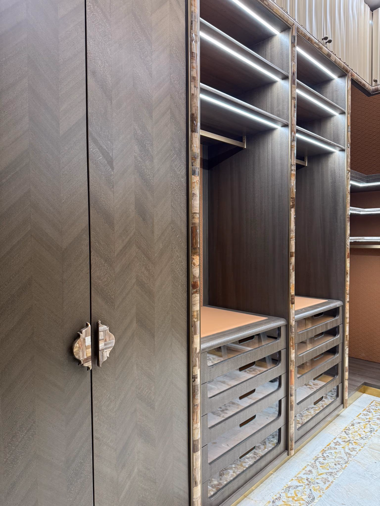 Dark walnut walk-in wardrobe with LED-lit open shelves, leather-topped pull-out trays, onyx-inlay pilasters and a chevron-veneer door with a stone handle.