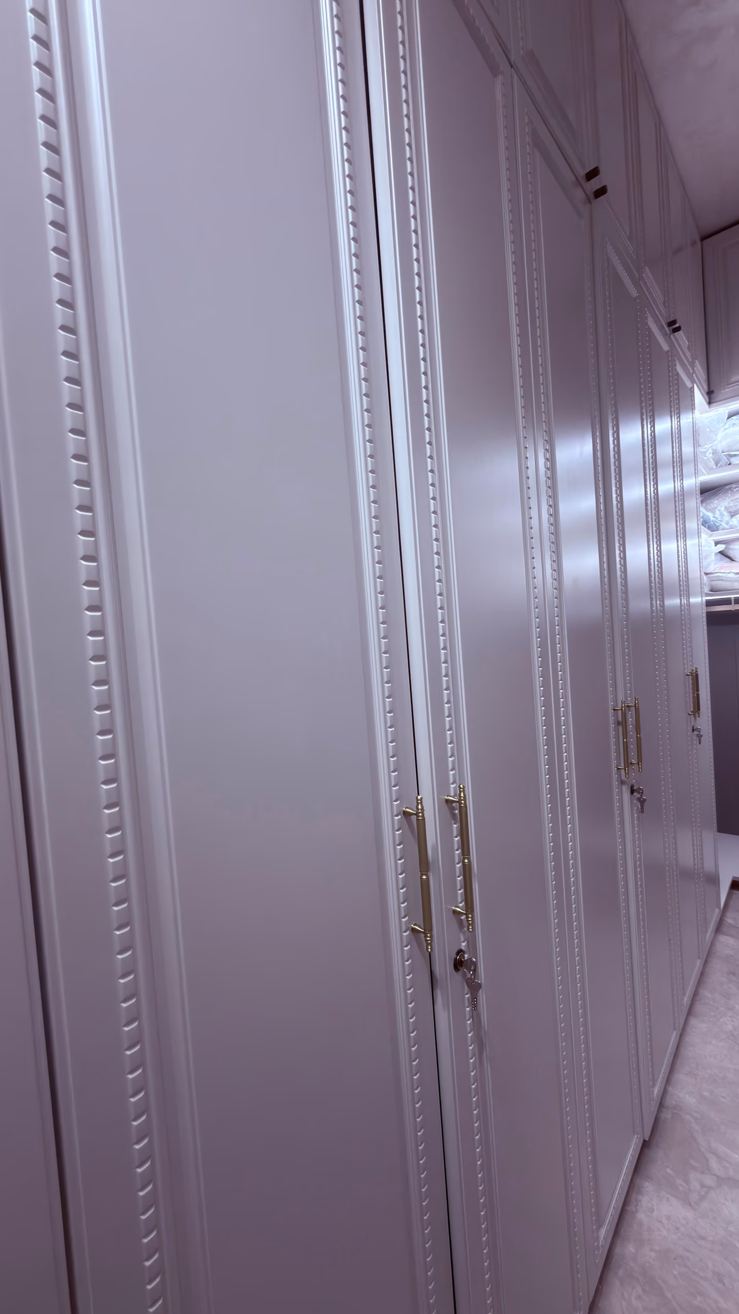 The soft-lilac beaded-trim wardrobe run seen down its length at an angle, its brass handles and open linen shelf catching light along a marble-floored corridor.