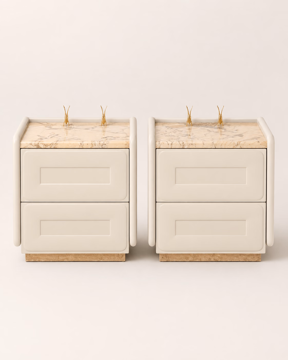 A pair of Designatore bedside tables shown alone on a bone ground, each with a cream Italian marble top in a raised gallery, two recessed-panel ivory drawer fronts, rounded column corners, slim gold pull handles and a solid marble plinth base.